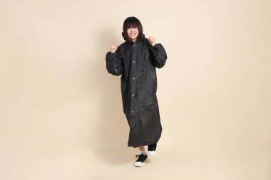 Siyah yağmurluk giyen genç bir Asyalı kadın ve evet, heyecanlı, bej arka planda izole edilmiş bir şekilde kutluyor. Yağmur mevsimi, hava durumu, sigorta kavramı.