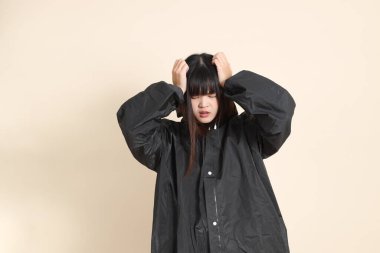 Siyah yağmurluk ve baş ağrısı giyen genç bir Asyalı kadın ya da bej arka planda izole edilmiş stres. Yağmur mevsimi, hava durumu, sigorta kavramı.