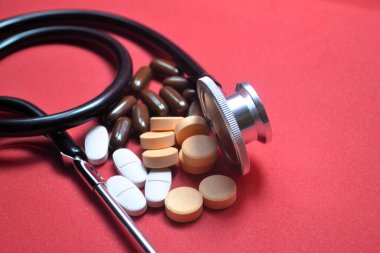 Sağlık hizmetleri markalaşması ve klinik eğitim görselleri için tıbbi tanıları ve ilaç çeşitliliğini vurgulayan kırmızı arka planda karışık haplar arasında stetoskop sarmalanmıştır.