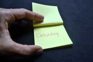 Koyu yüzey üzerine cumartesi mesajı içeren sarı nota. Haftasonu atmosferi, kişisel planlama ve verimlilik, ofis ve motivasyon için yaşam tarzı ritmi.