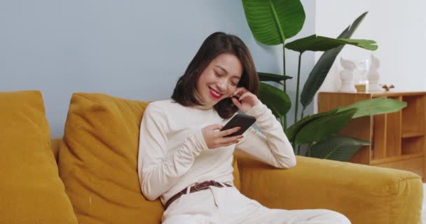 Красивая азиатская женщина сидит на диване с помощью мобильного телефона дома — стоковое видео