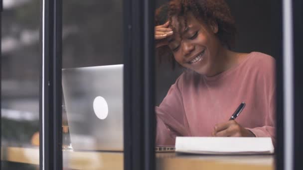 Молодая африканская девушка учится писать в кафе у окна — стоковое видео