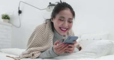 Genç Asyalı kız yatakta uzanıyor ve cep telefonuyla oynuyor.