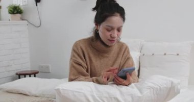 Genç kız cep telefonuyla yatakta oturuyor.