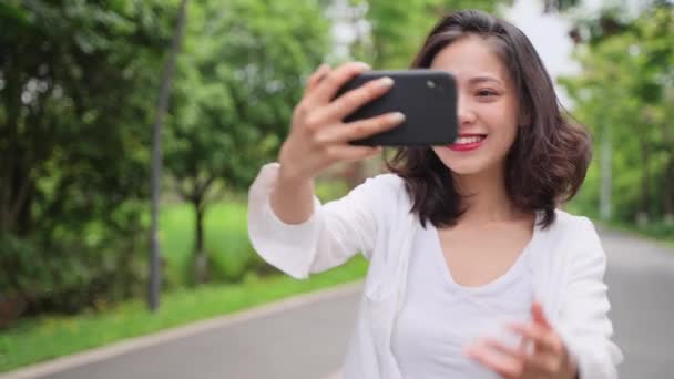携帯電話のビデオを見て夏の公園を歩くかなり若いアジアの女性のスローモーションチャット笑顔は喜びの女の子が道路を歩いてリラックス — ストック動画