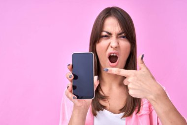 Esmer, mutlu, ağzı açık bir kız parmağı boş cep telefonu ekranını işaret ediyor ve pembe stüdyoda izole edilmiş fotokopi alanı var.