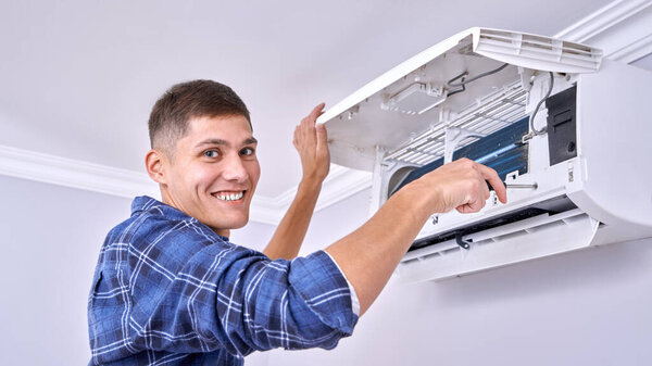 Caucasian male master in blue shirt cleans filters, installs and fixes air conditioner indoors