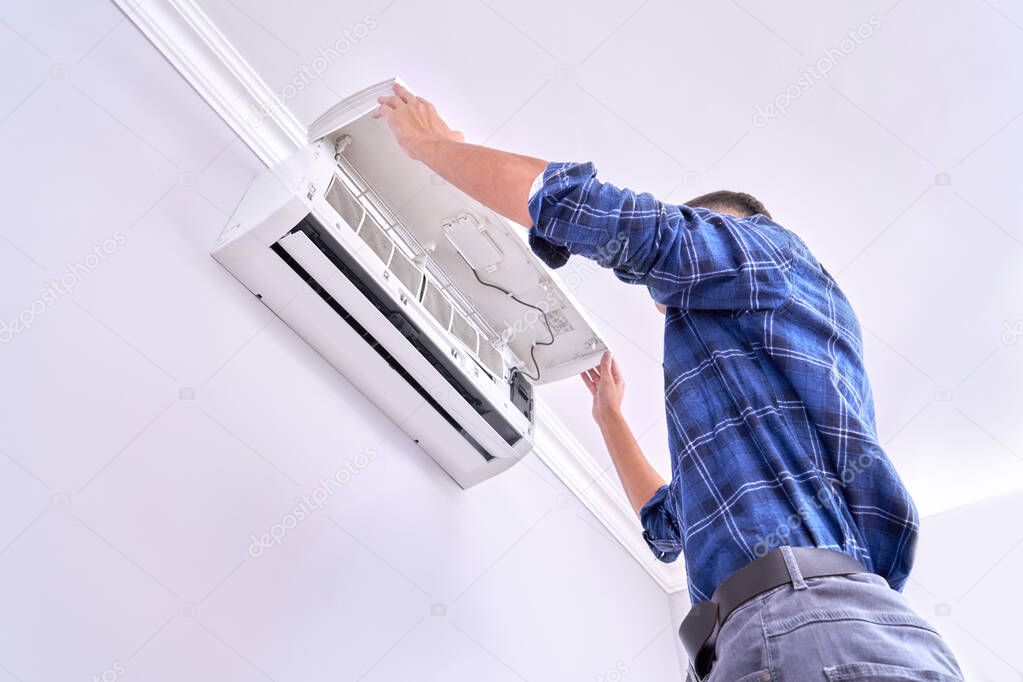 Caucasian male master in blue shirt cleans filters, installs and fixes air conditioner indoors