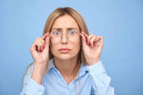 OMG! I don\'t believe you. Studio shot blonde girl with puzzled face and bulging eyes holds hands on the rim of glasses, emotional reaction to unexpected news isolated on blue background