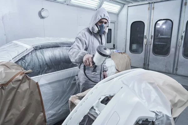 Car painting and automobile repair service. Auto mechanic in white overalls paints car with airbrush pulverizer in paint chambe