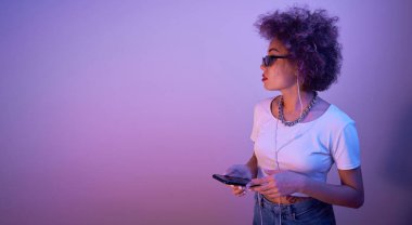Kıvırcık saçlı moda bir kız stüdyo arka planında akıllı telefon ve kulaklık kullanıyor.