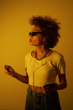 Kıvırcık saçlı moda kız sarı ışıkta kulaklıklı müzik dinler ve sever, hip hop tarzı dans eder ve dinlenir.
