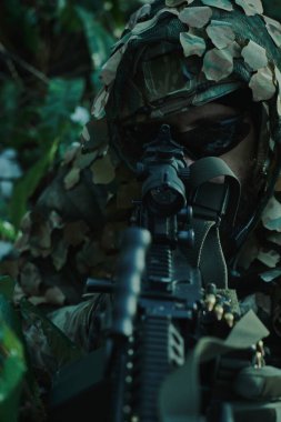 Profesyonel ekipmanlı, kasklı, havalı oyuncunun portresi ormanda silahla kurbana nişan alıyor. Savaşta silahlı bir asker