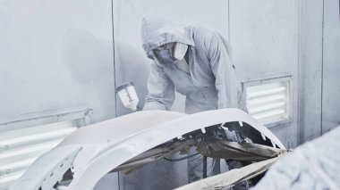 Araba boyama ve otomobil tamir servisi. Beyaz tulumlu oto tamircisi boya odasında boya fırçasıyla araba boyuyor.