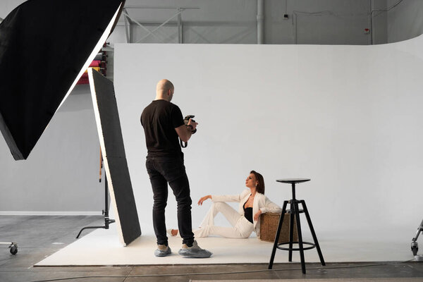 Fashion photography in a photo studio. Professional male photographer taking pictures of beautiful woman model on camera, backstage