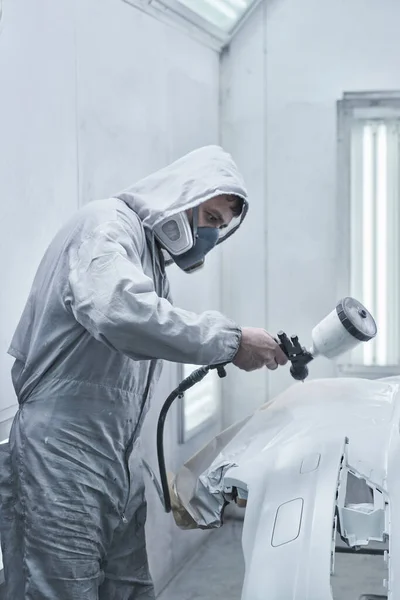 Car painting and automobile repair service. Auto mechanic in white overalls paints car with airbrush pulverizer in paint chambe
