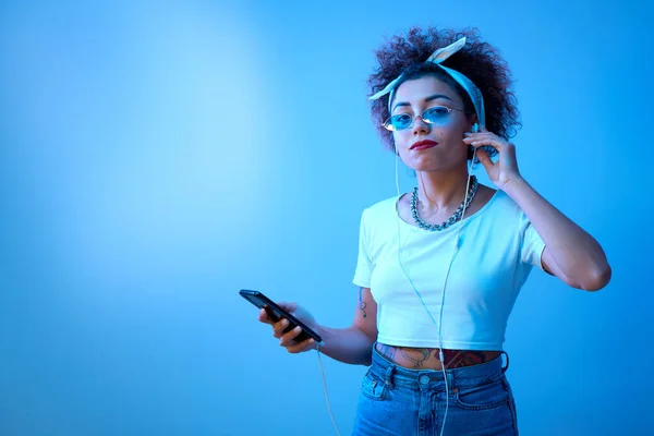Trendy girl with afro curls uses a smartphone and headphones on a studio background with copy space