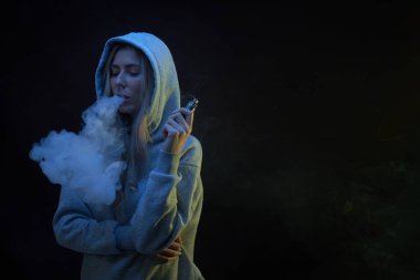 Gri kapüşonlu güzel sarışın kızın portresi siyah stüdyo arka planında izole edilmiş sigara içiyor, buhar dumanı bulutu, mini nargile.