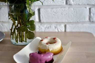 Two delicious donuts are sitting side by side on a pristine white plate placed on a table next to a lovely vase filled with beautiful flowers heats icing St. Valentines Day romantic present 