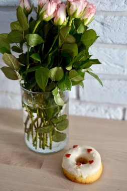 A delicious donut is sitting right next to a beautiful vase filled with colorful flowers on a lovely wooden table heats icing St. Valentines Day romantic present 