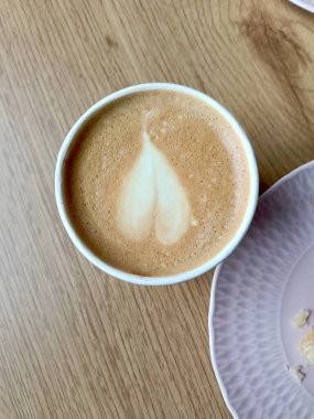 Kapalı köpüklü latte, ahşap sehpanın üzerinde düz döşeme, görülebilir pastel plaka kenarı, yumuşak gün ışığında kremsi doku, modern