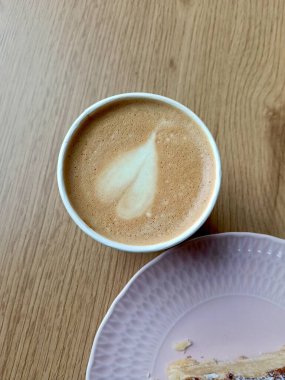 Köpüklü latte, hamur kırıntılı pembe tabak, sıcak ahşap masa yüzeyi, yumuşak sabah ışığı, samimi kahvaltı ritüeli.