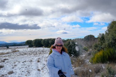 Açık renk ceketli ve yün şapkalı genç bir kadın karla kaplı tarlada yürür ve fotoğrafçıya bir kartopu atar..
