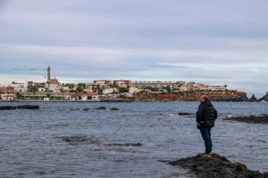Bir kayanın üzerinde, ayağında deniz olan bir adam, arka planda deniz feneri olan Cabo de Palos 'un sahil kasabasına bakar.