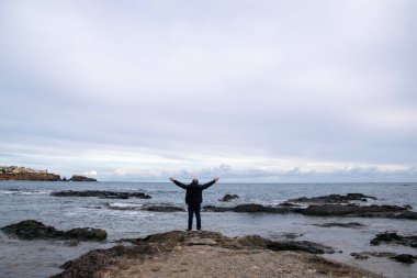 Bir adam, arka planda deniz olan beyaz duvarları ve bir ağacı olan bir ara sokakta yürüyor. Soğuk bir kış günü, gün batımında..