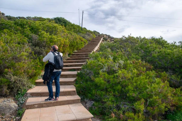 Kamerası için siyah bir sırt çantası taşıyan bir adam bitki örtüsüyle çevrili bir dağ merdivenine tırmanıyor..