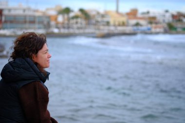 Bir kadın kış günü gün batımını düşünerek oturur, soğuk rüzgarın yüzüne vurmasıyla denize bakar..