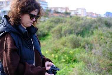 Kamerasıyla biraz zaman geçirdikten sonra, bir kadın, arka planda bitki örtüsü ve binaların olduğu bir alanda, sonucu görmek için ekrana bakar..