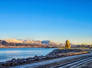 Arka planda karlı dağlar olan sakin fiyort suyunun yanında kayalık kıyıda duran parlak sarı deniz feneri. Yüksek kalite fotoğraf