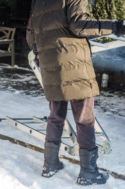 Sıcak ceketli ve botlu bir insan, güneşli bir kış gününde, açık hava işleri için karlı arka bahçede alüminyum bir merdiven taşır.