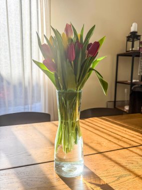 Cam vazodaki renkli bahar laleleri ahşap masadaki süs eşyası gibi. Sahne doğal ışıkla aydınlatılıyor ve sakin bir ev ortamı yaratıyor. - Evet. Yüksek kalite fotoğraf