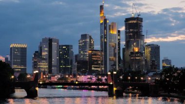 Şehrin ışıkları açıkken Frankfurt siluetinin ortasındaki göz kamaştırıcı mavi saat. Ana Nehir ön planda.