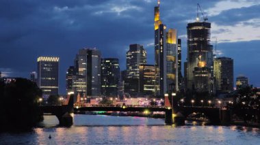 Karanlık çöktüğünde şehrin ışıkları açıkken Frankfurt siluetinin ortasındaki göz kamaştırıcı mavi saat. Ana Nehir ön planda.
