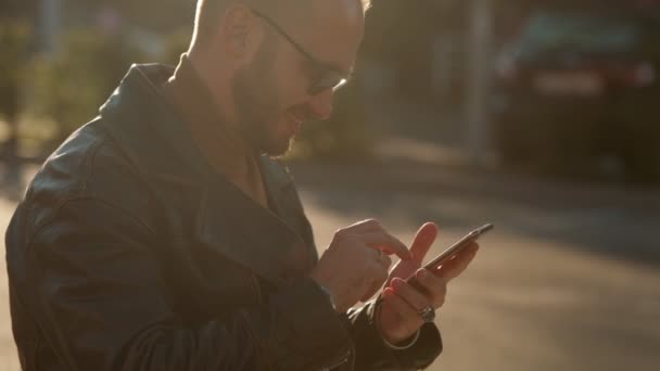 若い男が街の屋外の携帯電話で話す 革のジャケットを着たハンサムな白人男性 — ストック動画