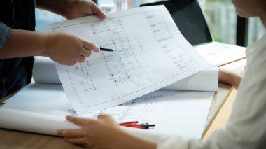 İki kişi bir binanın planına bakmaktalar. Bir tanesi planın bir kısmına işaret ediyor.