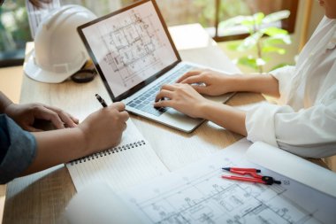 İki kişi bir dizüstü bilgisayar ve bir defter üzerinde çalışıyor. Bir kişi dizüstü bilgisayarda yazarken diğer kişi de deftere yazıyor. Dizüstü bilgisayar bir binanın çizimine açık.