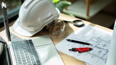 Masada dizüstü bilgisayar ve güvenlik gözlüğünün yanında beyaz bir şapka duruyor. Masanın üzerinde bir çift kırmızı ve siyah inşaat aleti de var.