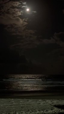 Ay, okyanusun üzerindeki karanlık gökyüzünde parlak bir şekilde parlıyor. Su sakin ve gökyüzü bulutlarla dolu. Sahne huzurlu ve sakin, ay ışığı sudan yansıyor.