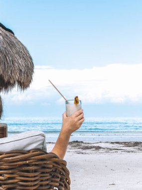 Hasır plaj sandalyesinde dinlenen bir kadın Holbox, Meksika 'da Karayip Denizi' ne bakan tropikal bir içecek tutuyor.