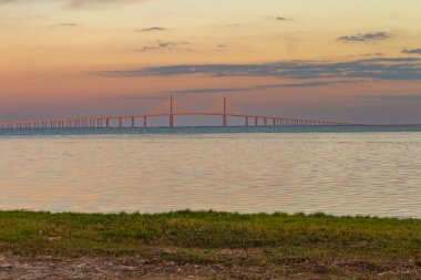 Sunshine Skyway Köprüsü şafak vakti Tampa Körfezi boyunca zarif bir şekilde uzanır. Yeni gün başlarken yumuşak altın ışıkla yıkanır. Sakin suda hafif dalgalanmalar sabah gökyüzünün canlı renklerini yansıtıyor, sakin ve huzurlu bir deniz manzarası yaratıyor..