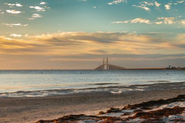 Işıl ışıl bir gündoğumu, parlak turuncu tonlarla gökyüzünü aydınlatan, Sunshine Skyway Köprüsü boyunca altın bir ışık yayar. Güneş ışınları sürüklenen bulutlara yayıldıkça köprü ufka karşı yumuşak bir siluete dönüşür..