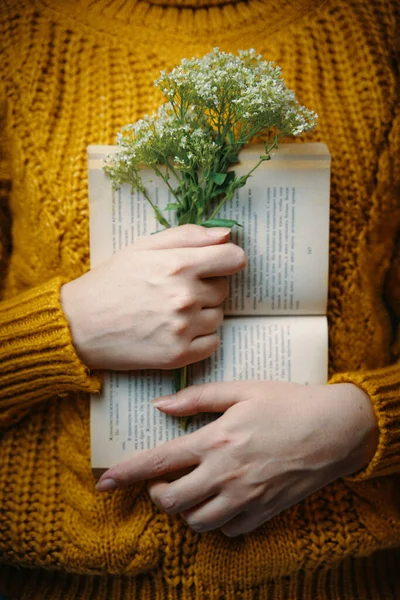 Female in sweater is holding book and bouquet.