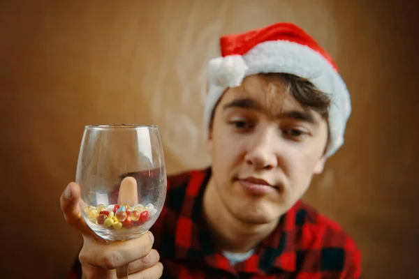 Guy in cappello di Babbo Natale tiene un bicchiere con varie pillole. — Foto Stock