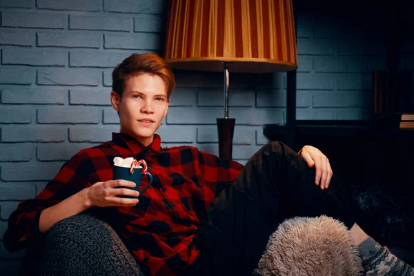 Young man in chair holds mug of hot chocolate with marshmallows. — Stock Photo, Image