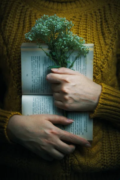 Female in sweater is holding book and bouquet.