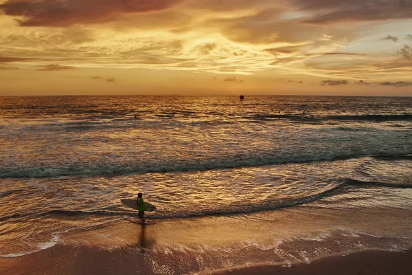 Garçon marchant le long de l'océan au coucher du soleil et portant dans ses mains une planche de surf — Photo